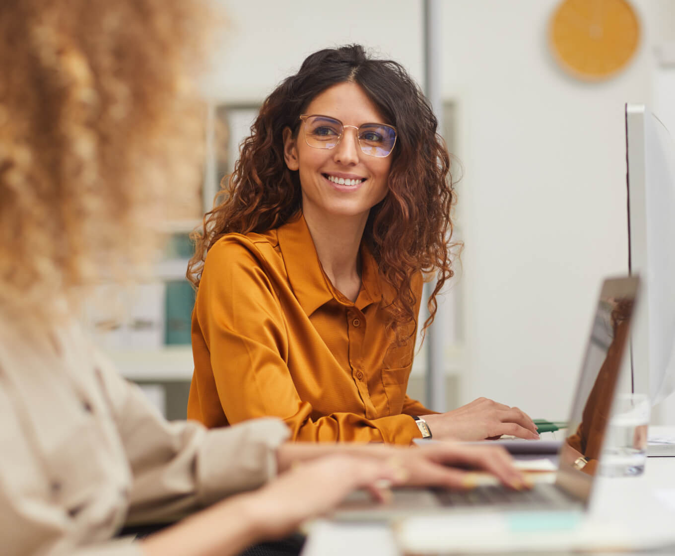 Two women sitting at a desk at work smiling at each other while on laptops