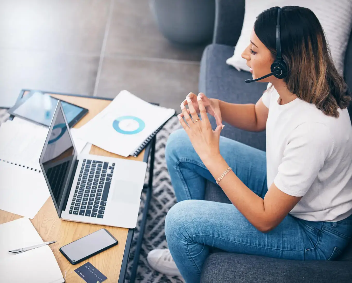 Woman sitting on couch talking to someone in her headset while looking at her laptop with papers all over table