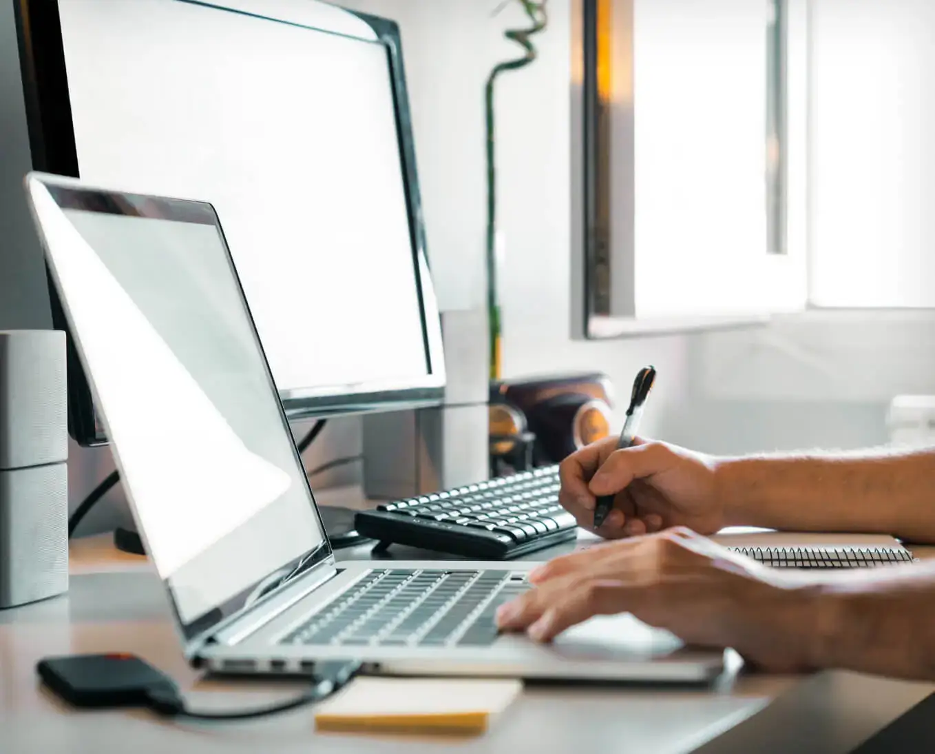 laptop and desktop screens with someone's hands typing and holding a pen